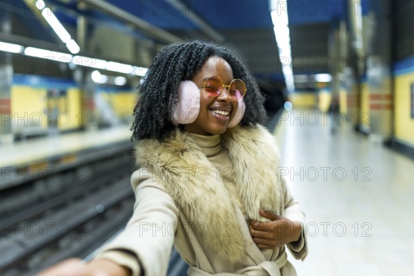 Young black woman wearing earmuffs and fur, taking a selfie with her smartphone in a subway station