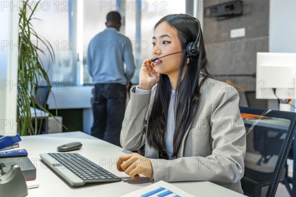 Young asian manager working diligently at a computer, wearing a headset in a sleek, modern