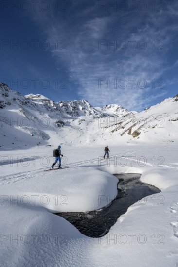 Small stream in a snow-covered mountain landscape, ski tourers in a valley, ascent to the