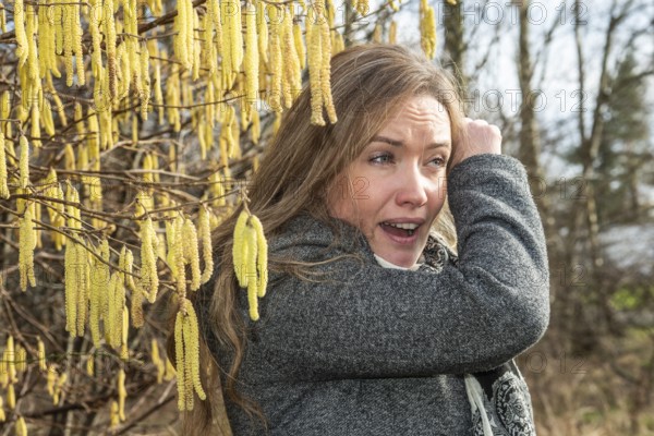 Young Caucasian woman with allergy problem stands by flowering hazel bush and sneezes in the crook