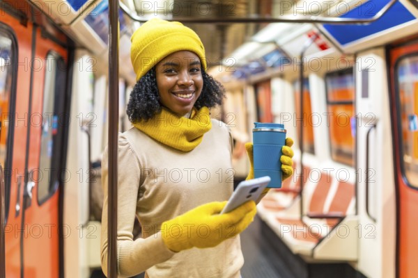 Smiling woman wearing yellow winter clothes using smartphone and drinking coffee inside subway