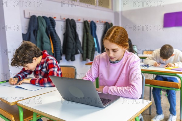 Elementary school students studying at individual desks, girl using laptop, boy writing in