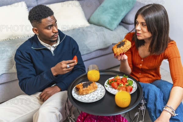 Young multiethnic couple eating pastries and strawberries with orange juice, enjoying breakfast at