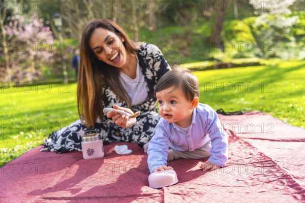 Happy mother feeding her baby solid food with a spoon, sitting on a blanket during a picnic in a
