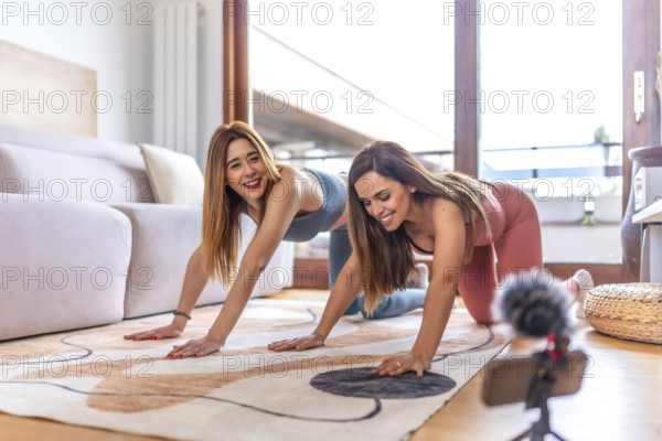 Two smiling women practicing yoga together in their living room, recording an online class with a