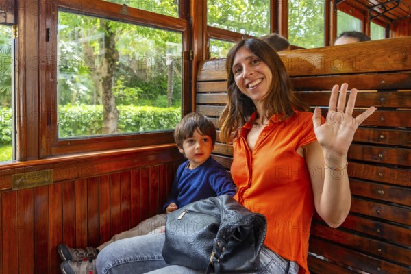 A woman and a child are sitting on a train. The woman is wearing an orange shirt and is waving at