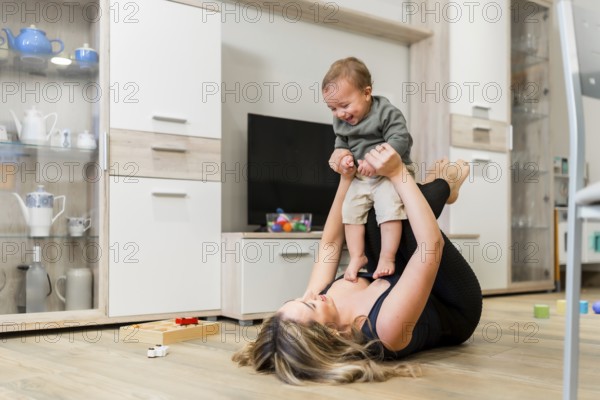 Young mother lying on the floor lifting her baby in the air while playing together in the living