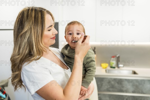 Loving mother feeding her baby son with a spoon in a bright modern kitchen, enjoying a tender