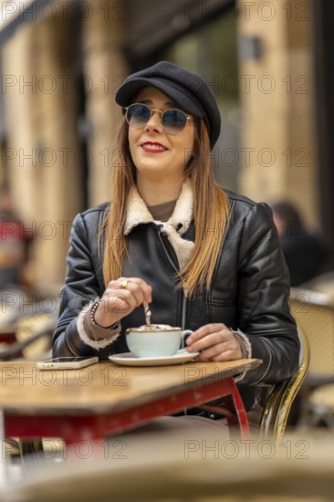 Young woman wearing sunglasses and hat stirring cappuccino at a parisian outdoor cafe, enjoying a