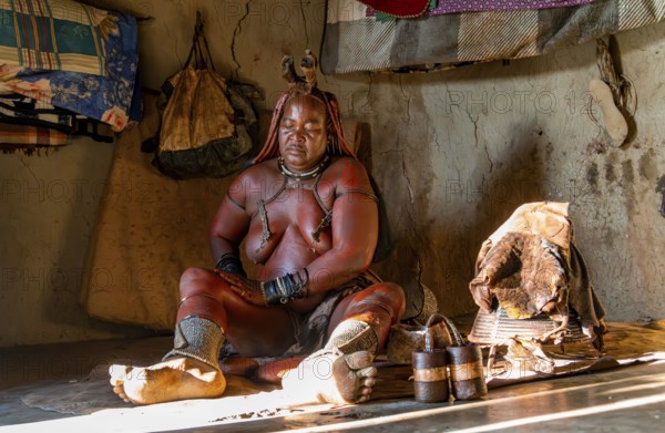 Traditional Himba woman applying ochre paint for body colouring, interior of the first woman's mud