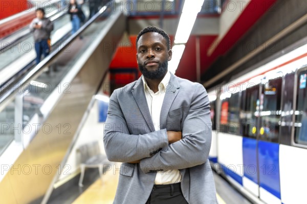 Young black businessman standing with crossed arms in subway station, waiting for transportation,