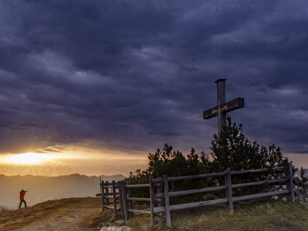 Photographer next to the summit cross on the Roßfeld in a dramatically overcast sky, the sun shines