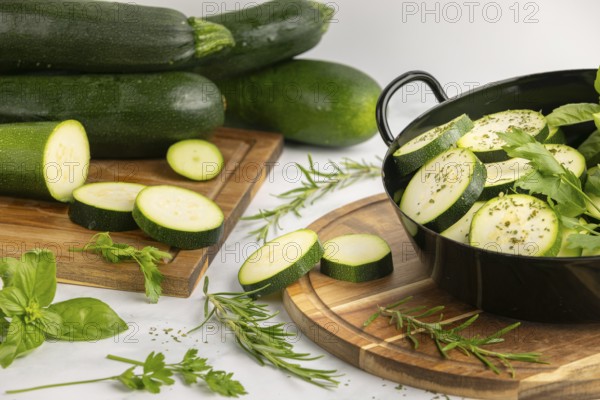 Sliced and seasoned courgettes with fresh herbs in a black pot, next to stacked courgettes on a