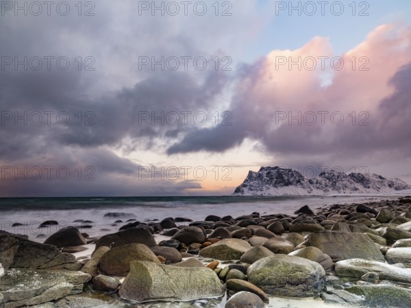 Rounded rocks on Utakleiv beach in a dramatic cloudy atmosphere, snow-capped mountains in the