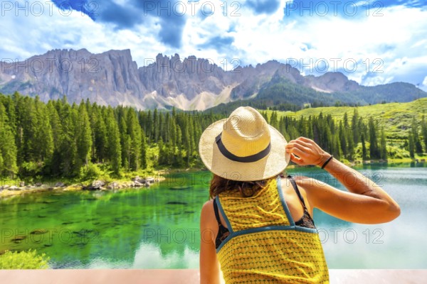 Woman wearing straw hat admiring lake carezza with latemar mountain range in background, dolomites,