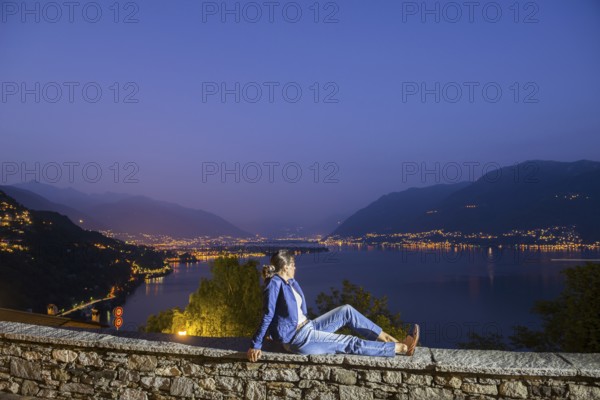 Woman Sitting on a Wall with Panoramic View over Lake Maggiore and Mountain in Dusk in Ronco sopra