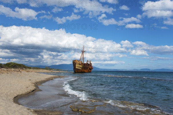 An abandoned, rusty shipwreck lies on a sunny beach with blue sky and clouds, Shipwreck Dimitrios,