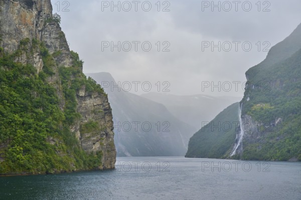 Geiranger Fjord with waterfall surrounded by rocks and mountains with overcast sky, Geiranger,