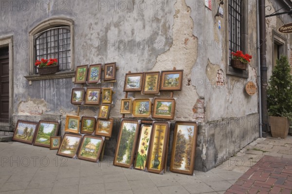 Framed paintings displayed against an old damaged grey cement wall in the Old Town Market Square,