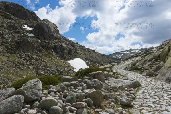 A stony path through a rocky mountain region with patches of snow under a cloudy sky, hiking area,