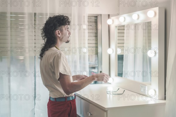 Young man with a mullet hairstyle standing in front of a lit mirror, holding items, dressed in a