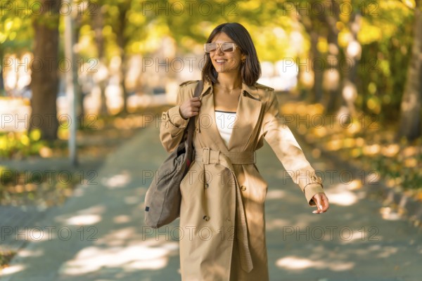 A woman wearing a tan trench coat and sunglasses walks down a sidewalk. She is carrying a brown