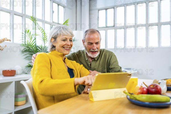 Happy senior couple using digital tablet at home pointing at the screen and smiling while eating