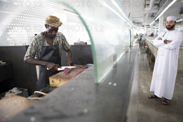 Omani man filleting a fish in Muttrah fish market, an Omani man in dishdasha watches, Muscat, Oman,
