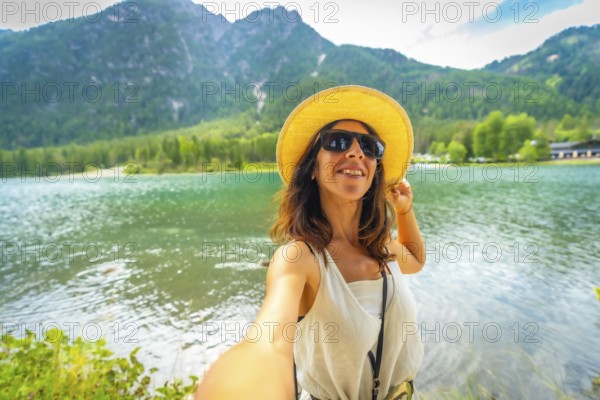 Tourist wearing sunglasses and a straw hat takes a selfie at lake dobbiaco with the dolomites in