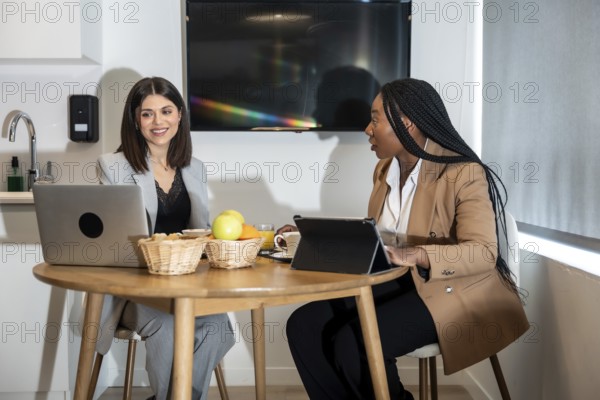 Two businesswomen are enjoying breakfast and working with laptop and tablet in their hotel room