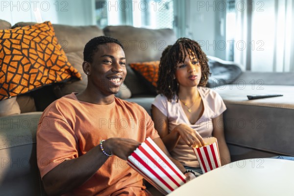 Young black couple enjoying a movie night at home, sitting on the sofa and eating popcorn from