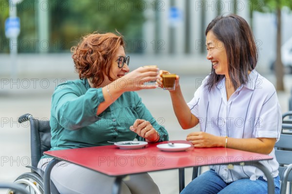 Woman with disability and asian friend toasting with coffee sitting on an outdoor cafeteria in the