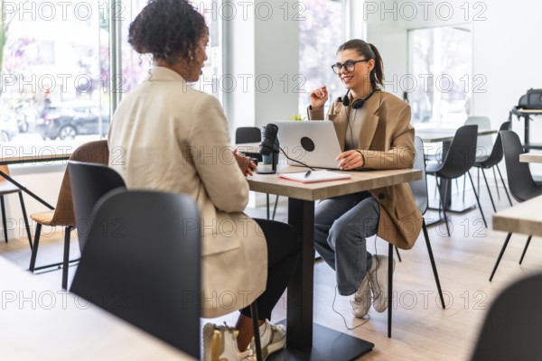 Two women engaging in a podcast recording session at a cafe, utilizing a laptop and microphone for