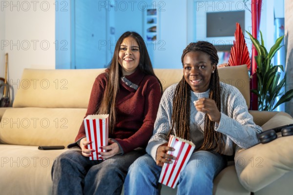 Two happy teenage girls eating popcorn while watching a movie at home, sharing a moment of leisure