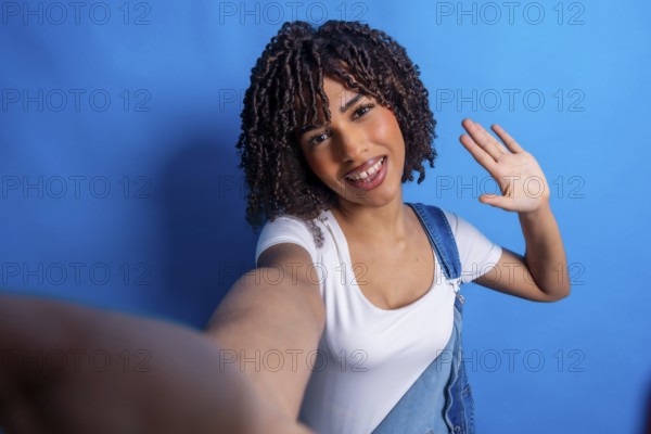Smiling young woman with curly hair, taking a selfie and waving her hand against a vibrant blue
