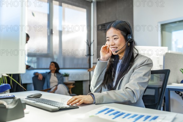 Young asian manager wearing headset working with computer in a coworking office, colleagues talking