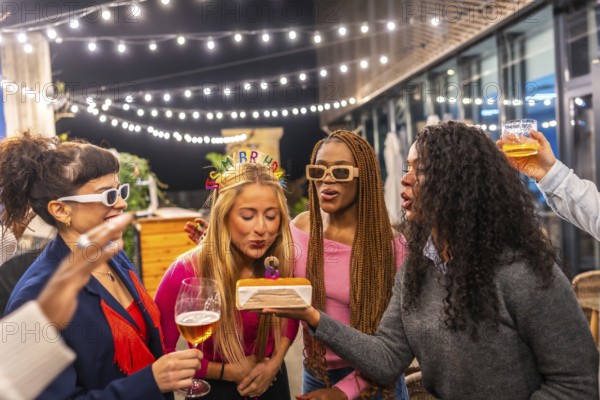 Blonde woman blowing candles of birthday cake celebrating party with friends at night in the