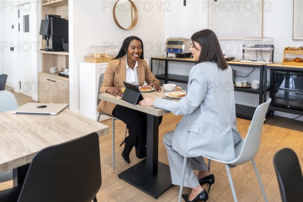 Two businesswomen are enjoying breakfast and discussing work using a digital tablet in a hotel