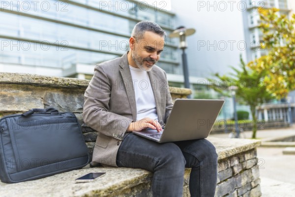 Happy mature caucasian businessman using laptop sitting on a stone bench outdoors in the financial