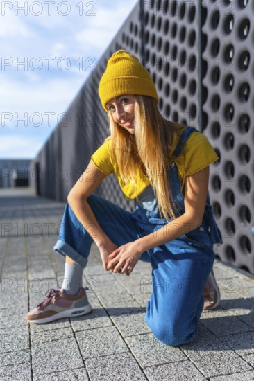 Blonde dancer wearing yellow beanie, yellow t shirt and blue dungarees, kneeling and posing in a