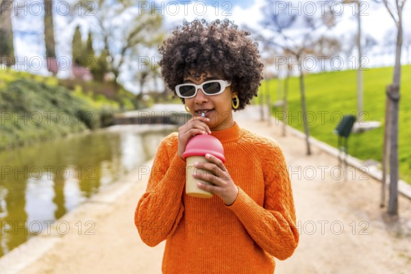 Stylish young woman with an afro hairstyle sips a beverage through a straw in a serene park setting