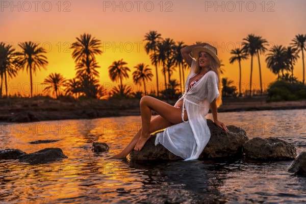 Full length photo of a caucasian woman sitting sensual on water's edge during sunset on a tropical