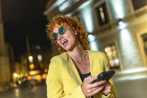 Modern woman in colorful casual clothes and blue sunglasses using smartphone laughing in the city