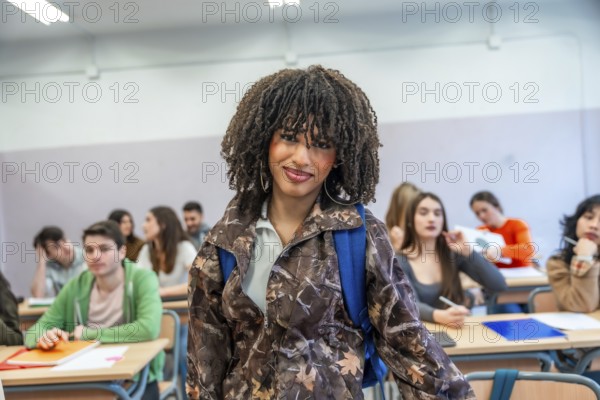 Confident young university student standing in classroom with backpack, classmates studying in
