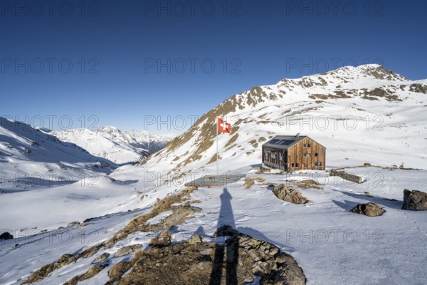 Swiss flag and mountain hut Keschhütte in front of mountain landscape in winter, behind summit of