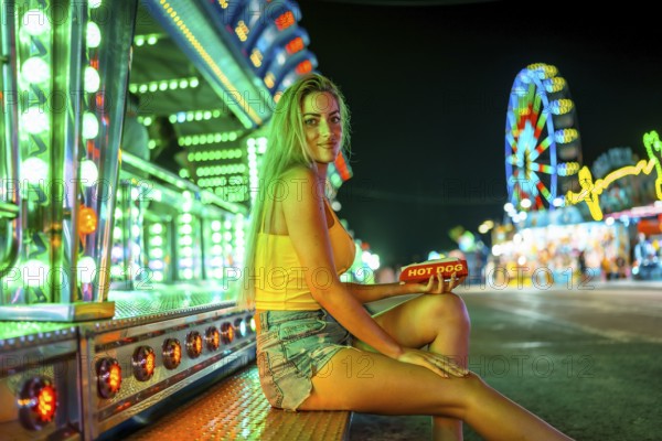 Blonde woman sitting alone about to eat a hot dog in a fair at night