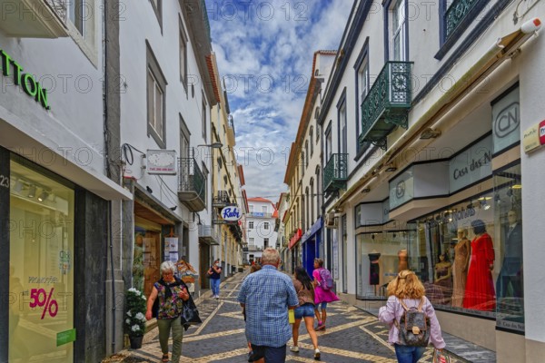 People walk through a busy shopping street with modern shops and historic buildings under a bright