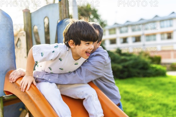 Grandfather holding grandson sliding down an orange slide at playground, having fun together on a
