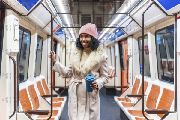 Smiling young black woman wearing winter coat and pink woolen hat holding reusable coffee cup