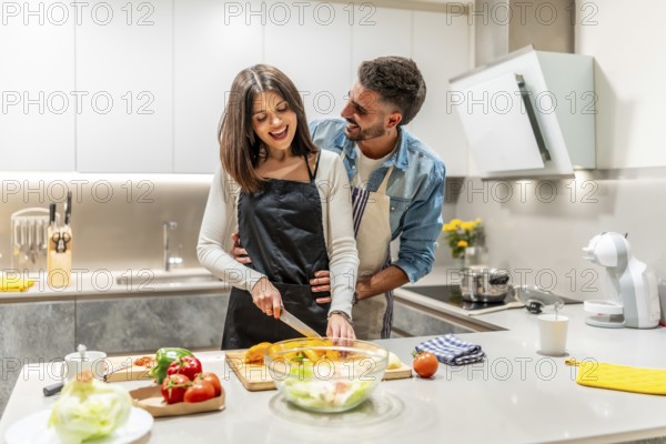 Young smiling couple wearing aprons preparing a healthy vegetable salad together in their modern